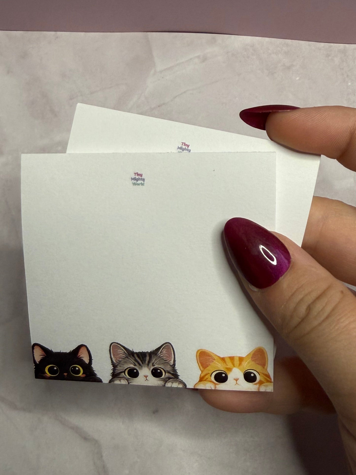A hand with dark red nail polish holds a stack of white notepad sheets. The top sheet features cute cartoon cat illustrations peering over the edge, including orange, gray, and black cats.