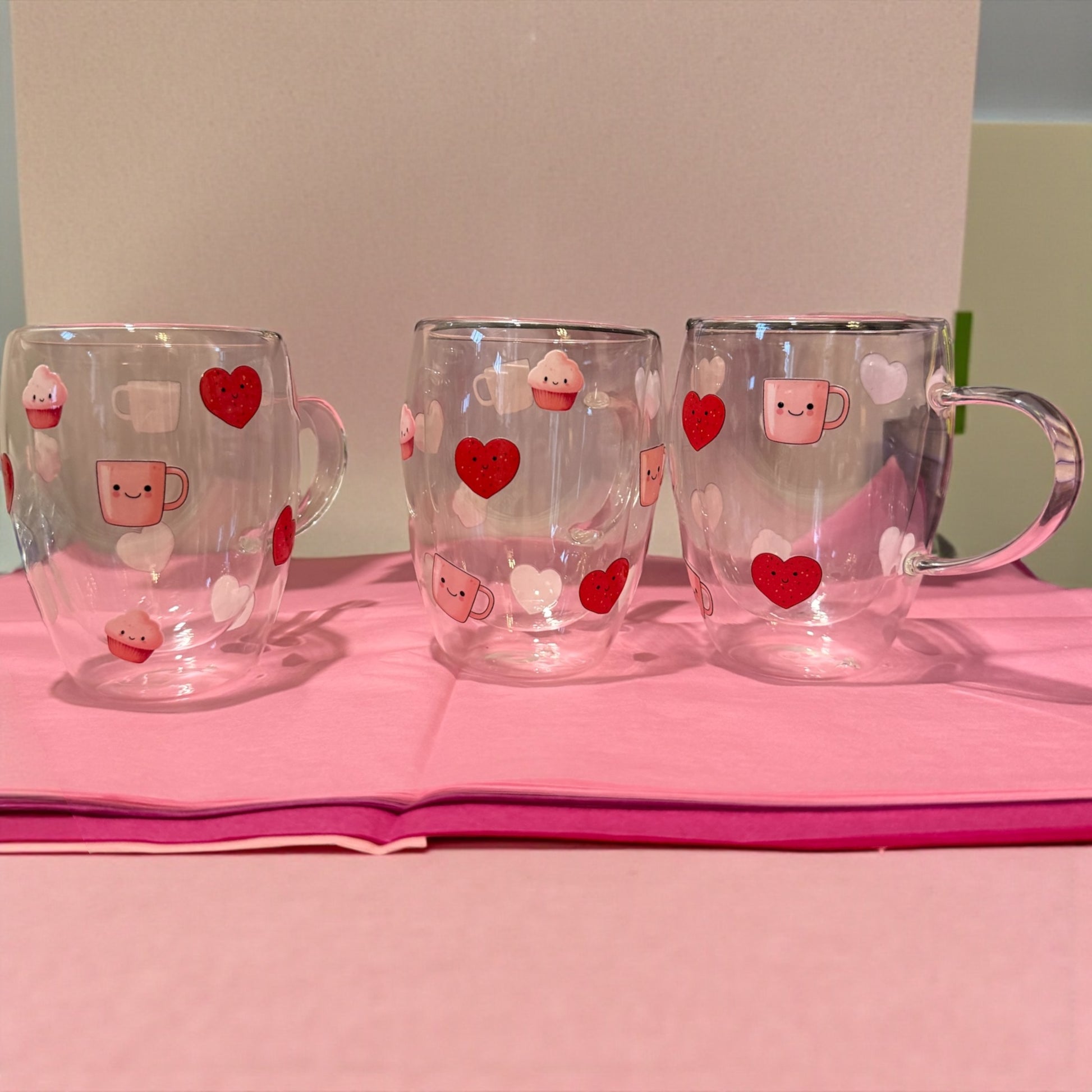 Three clear mugs with heart and cupcake designs on a pink surface.