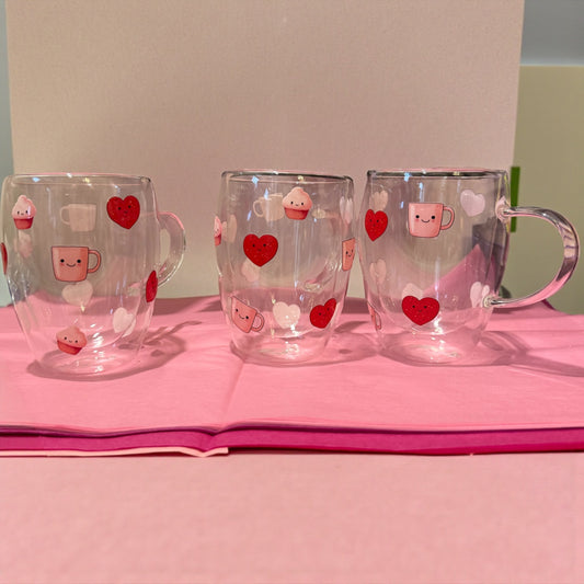 Three clear mugs with heart and cupcake designs on a pink surface.