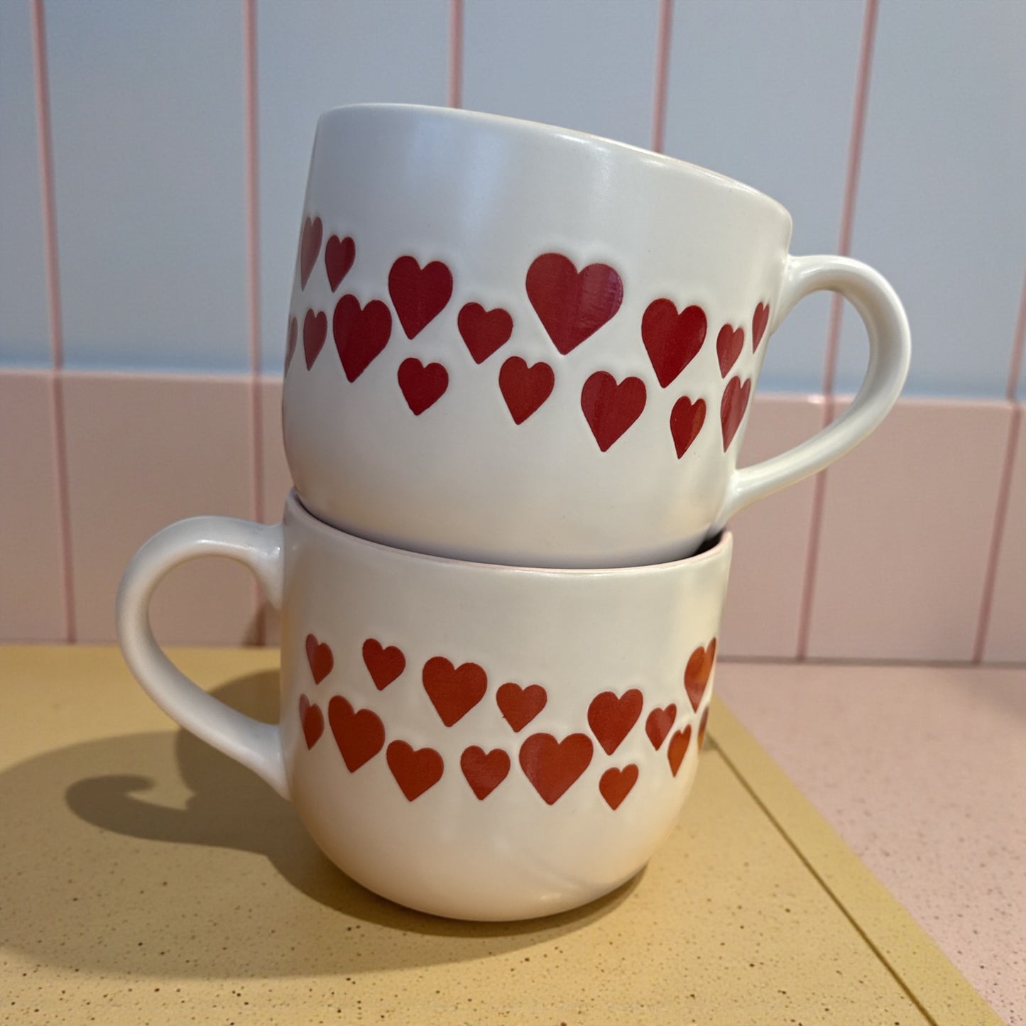Two white mugs with red heart patterns stacked on a countertop.